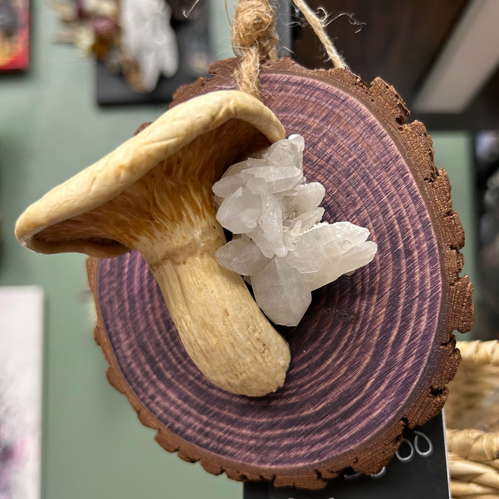 Photo of mushroom and crystal on wood ornament. The mushroom is tan and is next to a white crystal. Both are mounted on a wood cross-section of a small tree. It is stained purple, but you can see the tree rings through the stain.
